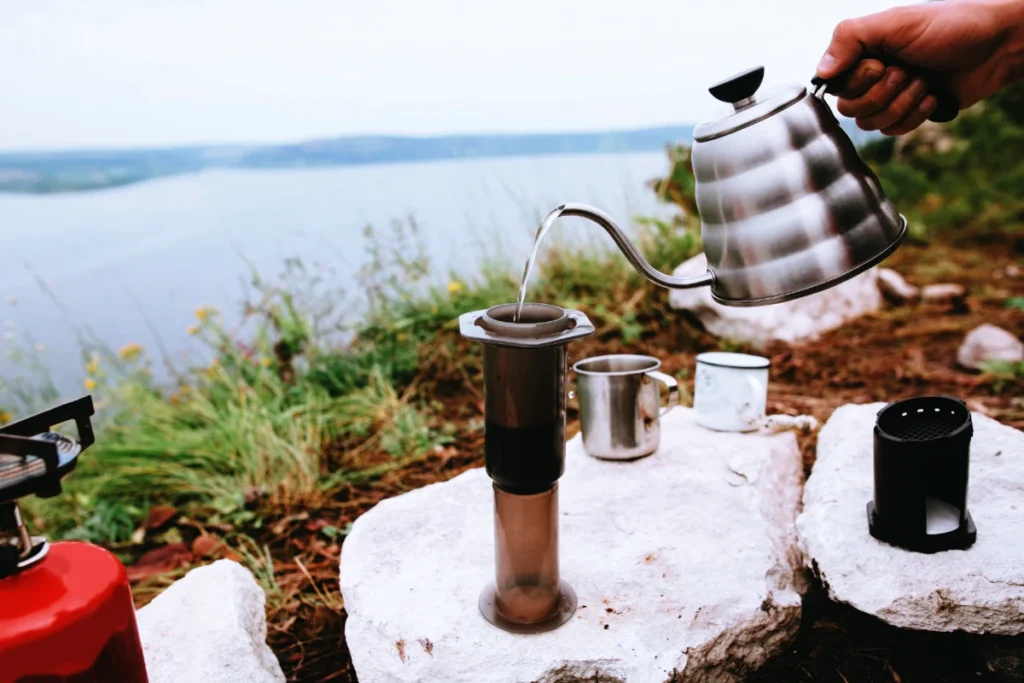 a photo of a person making camp coffee with an aeorpress on a hill overlooking a lake. #campingcoffee