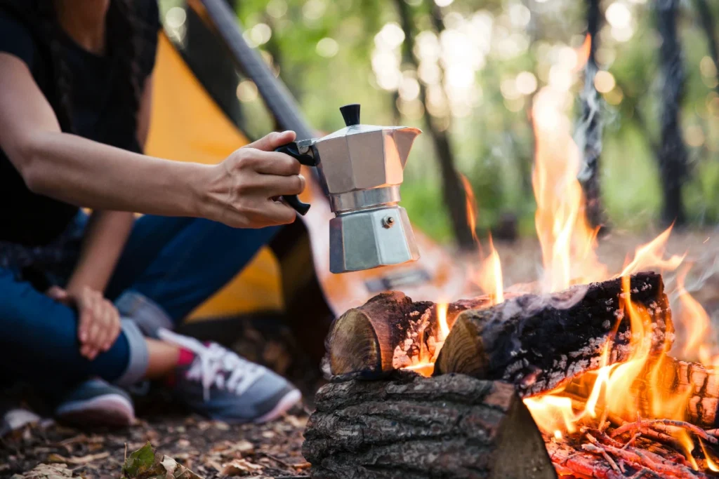 a person making camp coffee holding a moka pot over a campfire in nature with a tent in the background